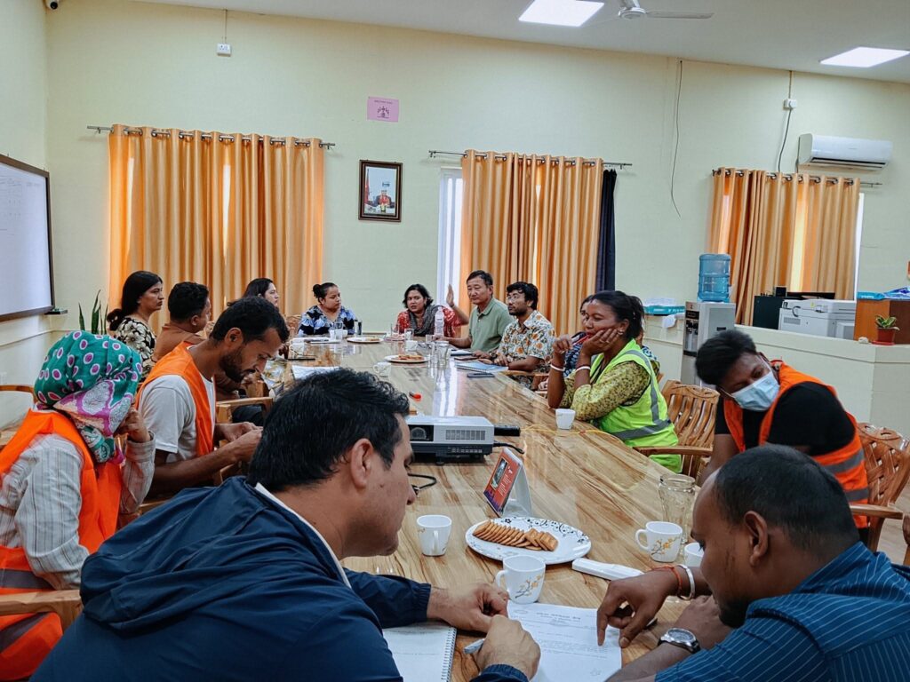 A group of people sitting around a large table in a conference room. Surendra Dhimal and Baldev Singh Gomden are visible. They are engaged in a discussion, likely about PSEAH prevention. The recipients are local governance and civil society members from Mechinagar Municipality, Jhapa. The setting is a conference room with yellow walls and orange curtains. The mood appears to be collaborative and informative.
